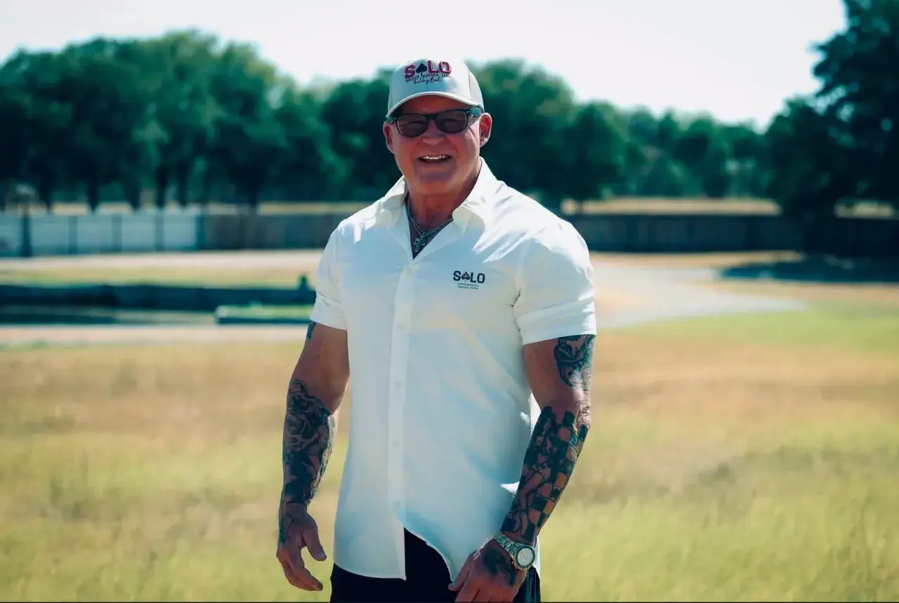 A smiling man with arm tattoos wearing a white short-sleeve shirt, cap, and sunglasses stands outdoors in a grassy field with trees in the background.