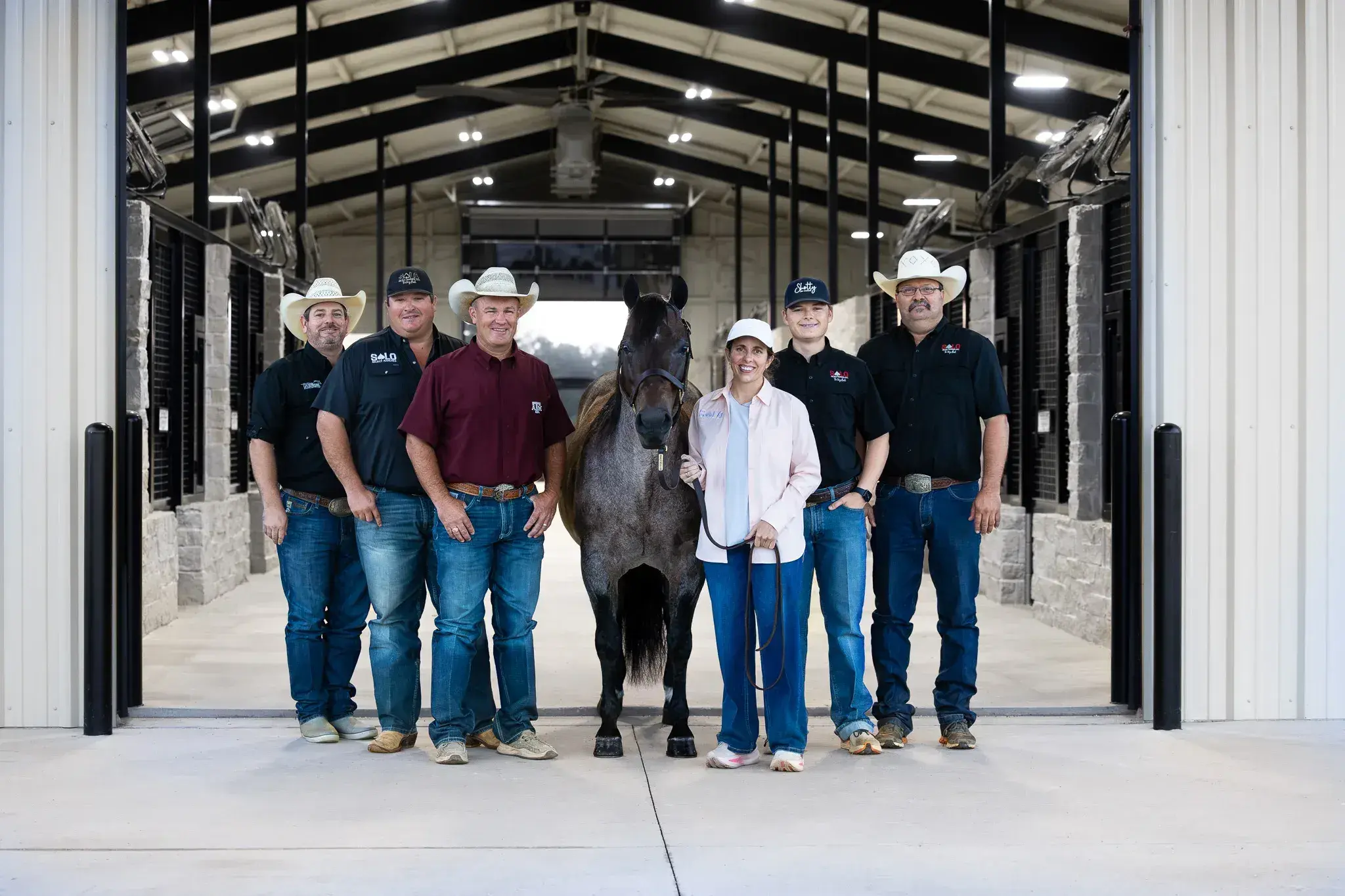 Six people, five in cowboy hats or caps and jeans, stand grouped around a roan horse in the open concrete aisle of a metal barn.