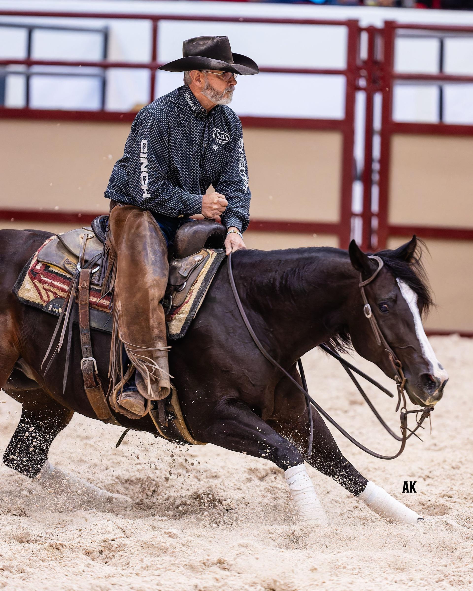 A dark cutting horse with a white blaze deep in a sliding stop under a rider in a black hat, sand flying