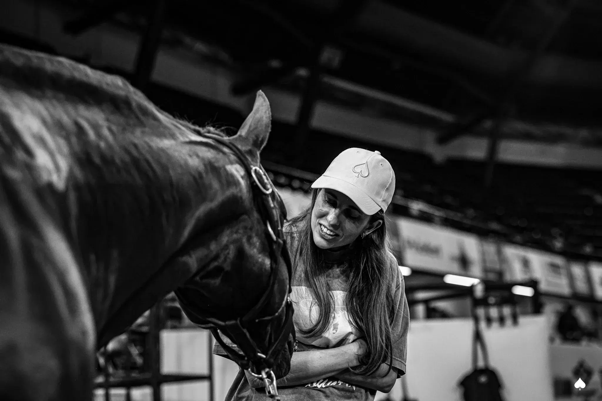 A black-and-white photo of a smiling woman wearing a cap and t-shirt standing beside a dark horse in a halter inside an indoor arena.