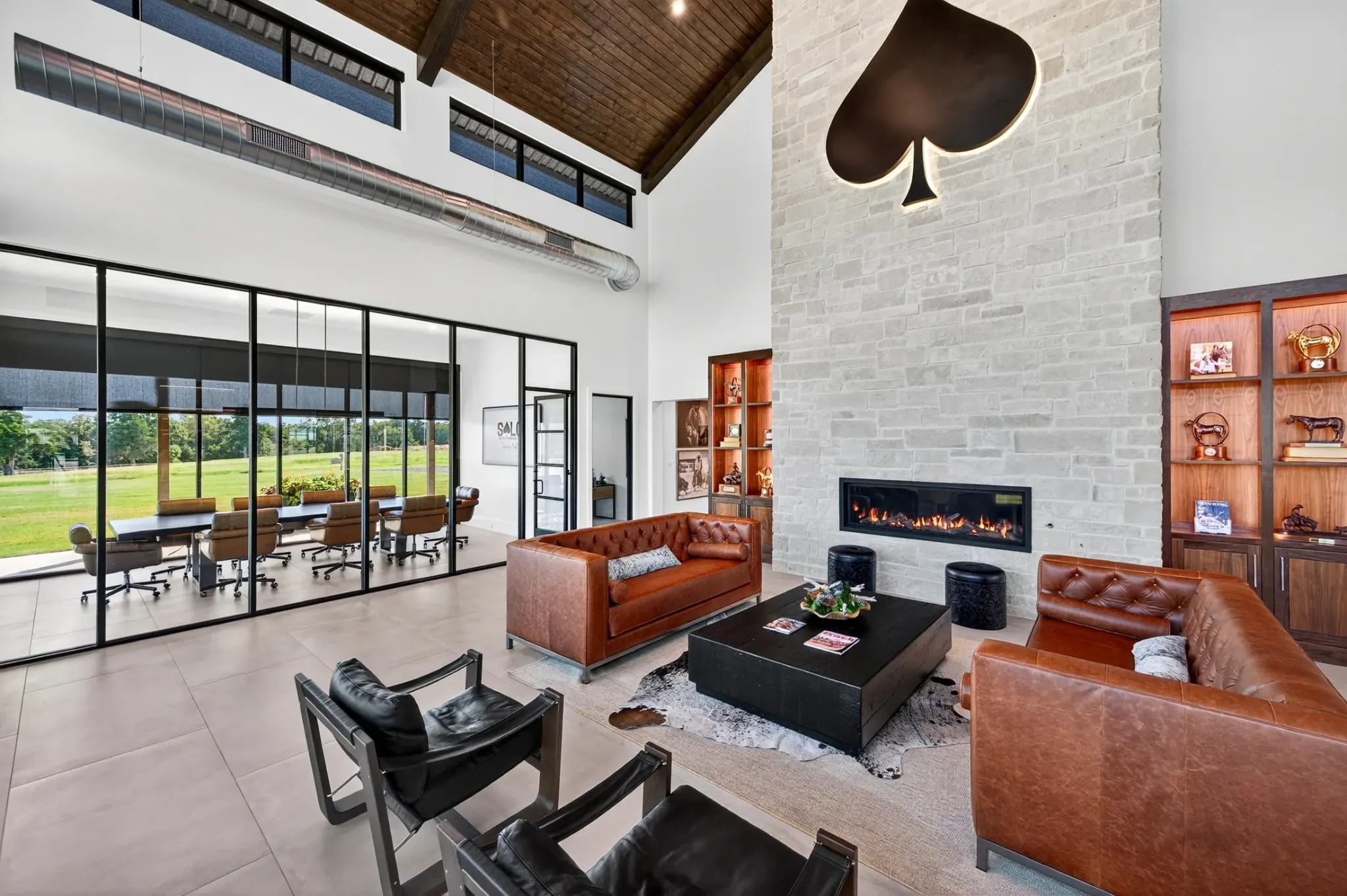 A modern high-ceilinged interior lounge with brown leather sofas, black leather chairs, a stone fireplace with a large spade emblem above it, and glass walls overlooking a green field.