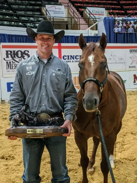 A man in a black cowboy hat and gray jacket stands holding a bronze trophy and belt buckle next to a chestnut horse with a small star and a halter in an indoor arena with sponsor banners and empty seating in the background.