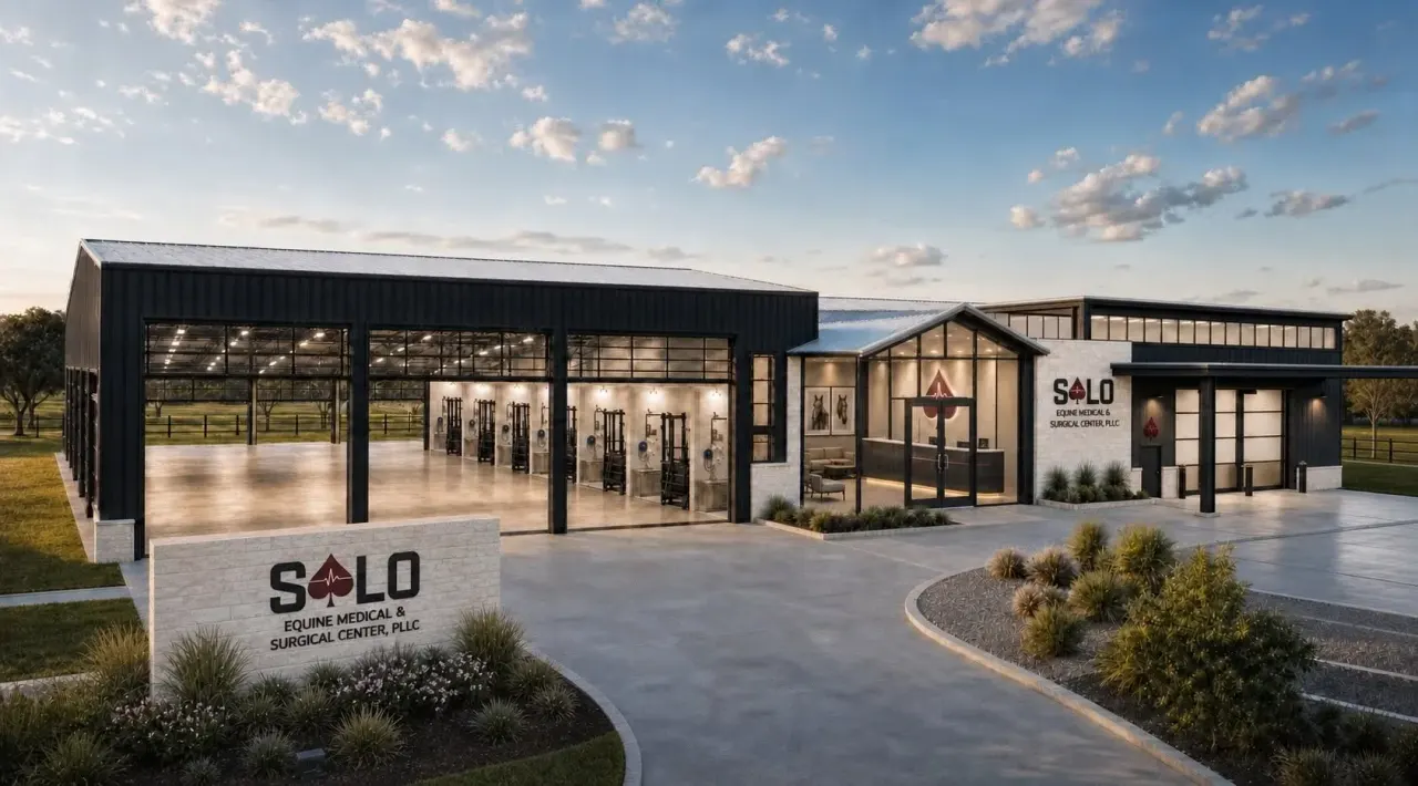 A modern black metal equine medical facility with large glass doors, lit interior stalls, a landscaped entrance sign, and concrete driveway under a partly cloudy sky at dusk.