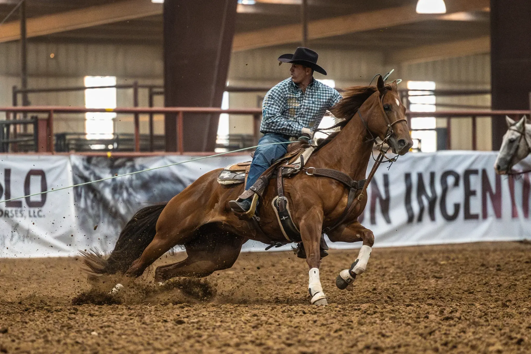 A rider in a black hat and blue plaid shirt sits a sorrel horse with a blaze and white hind leg markings as it slides and turns in the dirt of an indoor arena, with a gray horse visible in the background.