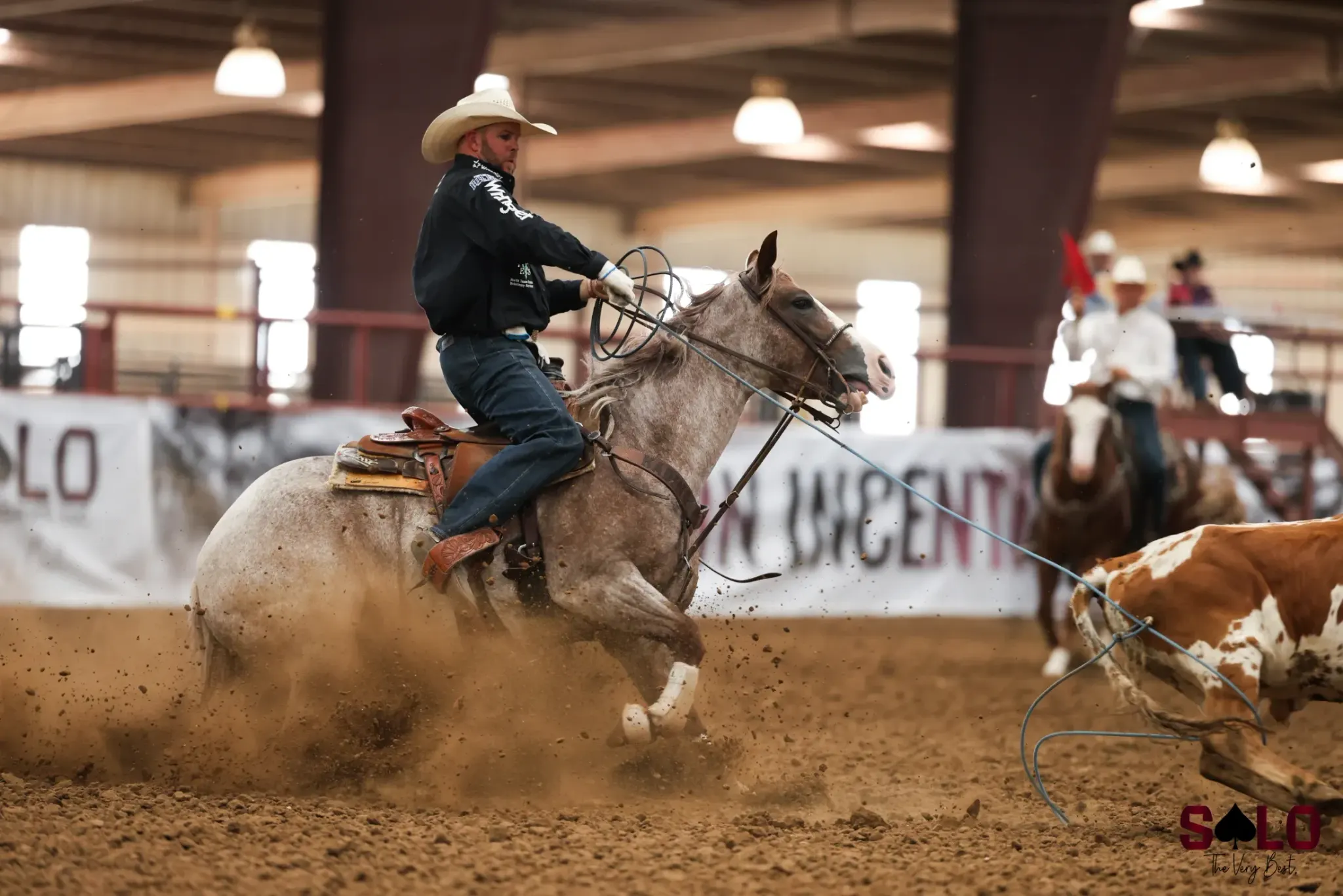 A rider in a white cowboy hat and dark shirt rides a red roan horse with a rope in hand, chasing a brown and white steer across a dirt indoor arena while mounted judges watch in the background.