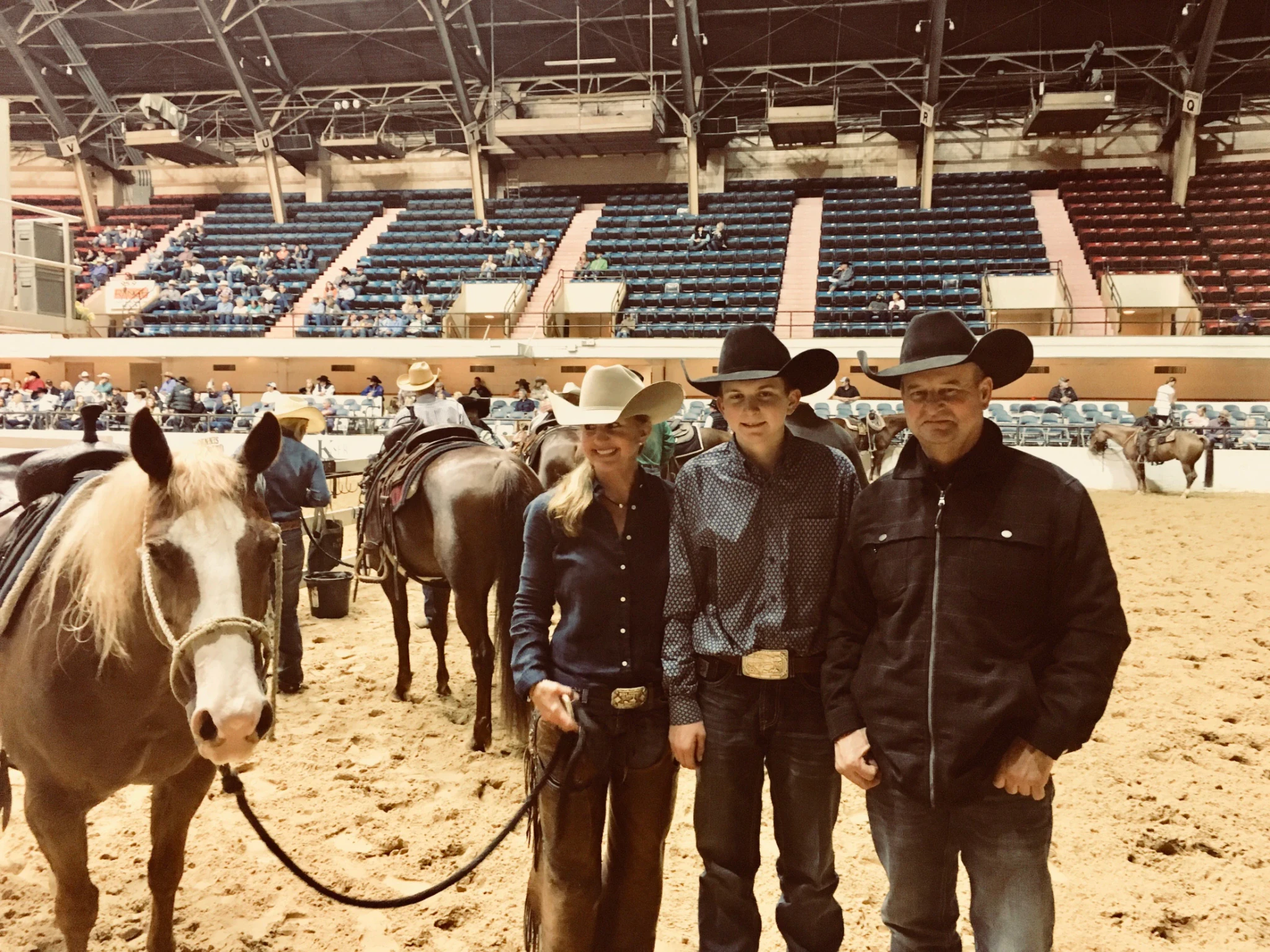 Three people in cowboy hats stand together in an indoor arena next to a palomino horse with a blaze, with other horses and spectators in the stands behind them.