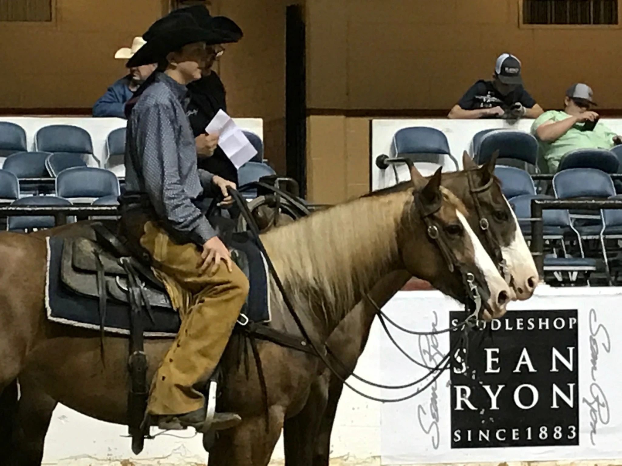 A rider in a black cowboy hat and tan chaps sits a western saddle on a chestnut horse with a blaze near empty blue arena seating, where two people watch and another person stands holding a paper.