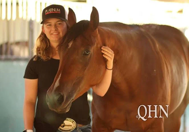 A woman in a black shirt and cap stands beside a bay horse, resting her hand on its neck in a stable area.