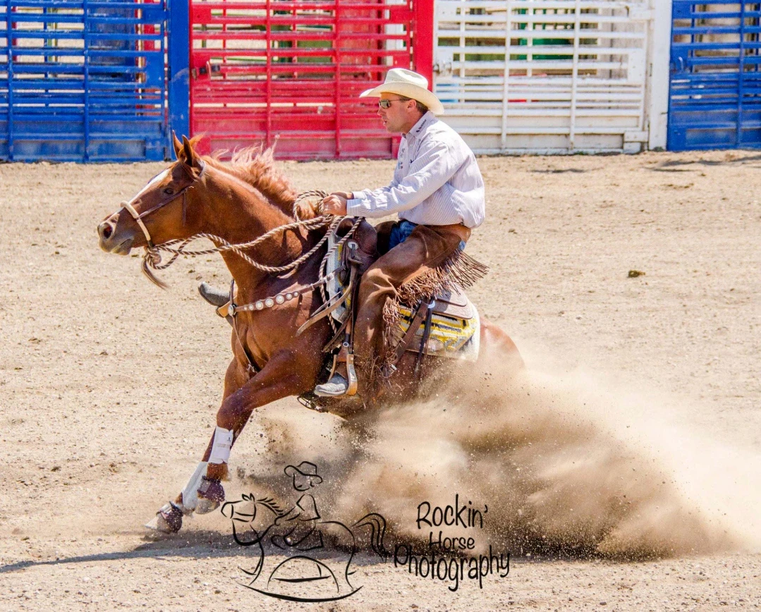 A chestnut horse with a blaze and white front leg markings performs a sliding stop in a dirt arena while a rider in a light shirt, chaps, and white cowboy hat sits the maneuver, kicking up a cloud of dust.